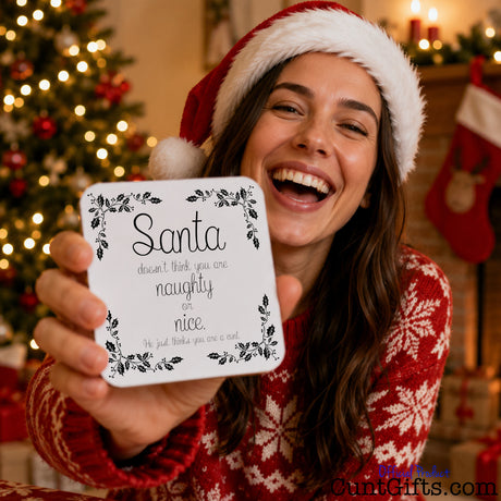 You are not naughty or nice you're a cunt - Christmas Drinks Coaster held by laughing brunette woman in Christmas hat and jumper in festive scene with Christman tree and lights.