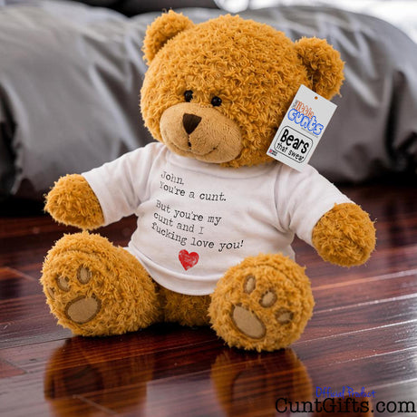 A brown plush personalised teddy bear wearing a white graphic T-shirt saying You're a Cunt and I Fucking Love You. The bear sits on a wooden floor in front of a grey bed. 
