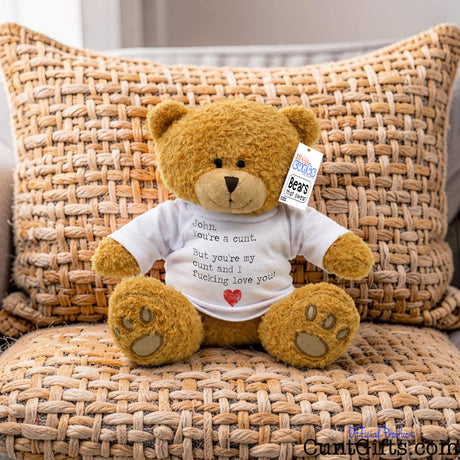 A brown teddy bear plush toy sitting on a woven chair, wearing a white T-shirtthat says "You're a Cunt and I Fucking Love You" -  and a small red heart. A hangtag is visible on the right side of its head.